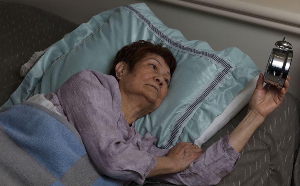 Ældre kvinde ligger i seng med vækkeur i hånden