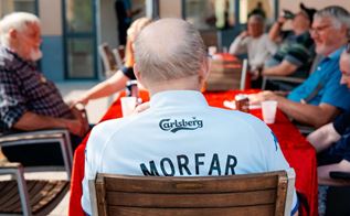Beboere på et plejehjem sidder rundt om et bord, og centralt placeret ser man en mand med måne bagfra iført en fødboldtrøje, hvor der står 'morfar' på ryggen. 