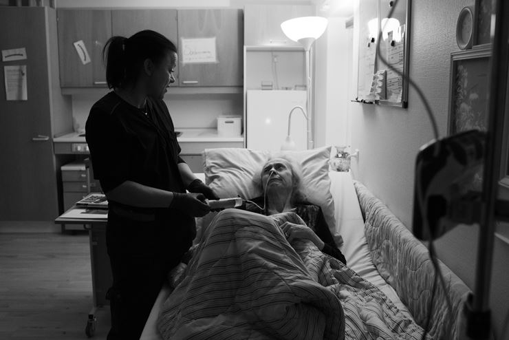 En sosu-assistent står ved siden af en kvindelig beboers seng og giver hende mad gennem sonde. Den ældre kvindelige beboer ligger i sin seng og kigger på sosu-assistenten. 